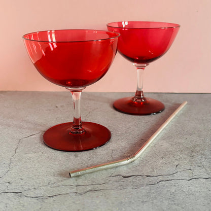 Two red translucent stemmed cocktail glasses on a gray concrete surface with a pink background, alongside a clear glass straw.