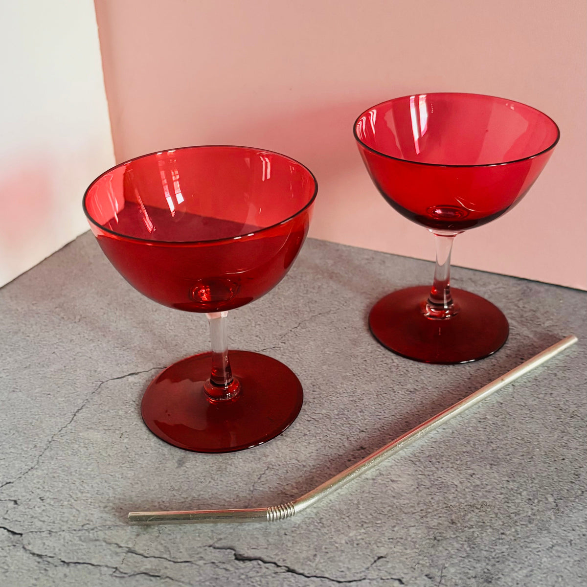 Two red translucent cocktail coupe glasses on a gray textured surface with a pink background wall.