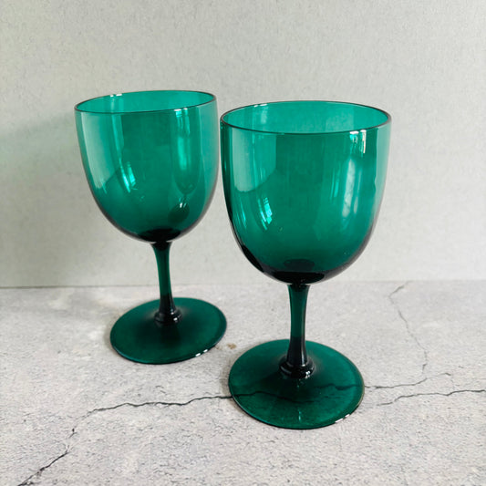 Two translucent emerald green wine glasses with round bases, standing on a light gray textured surface against a neutral light gray background.