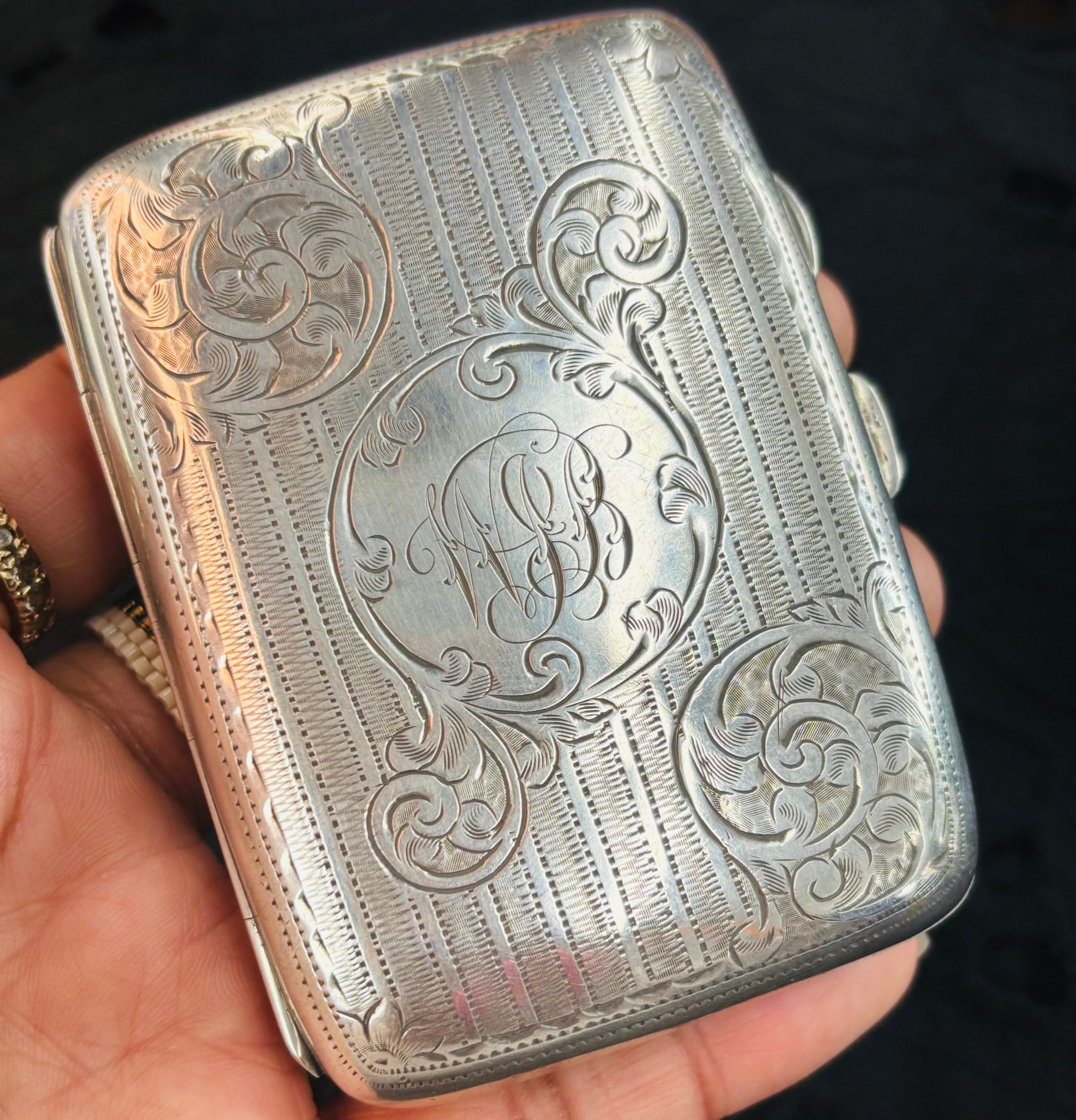 Silver cigarette case with intricate engravings held in a hand against a dark background