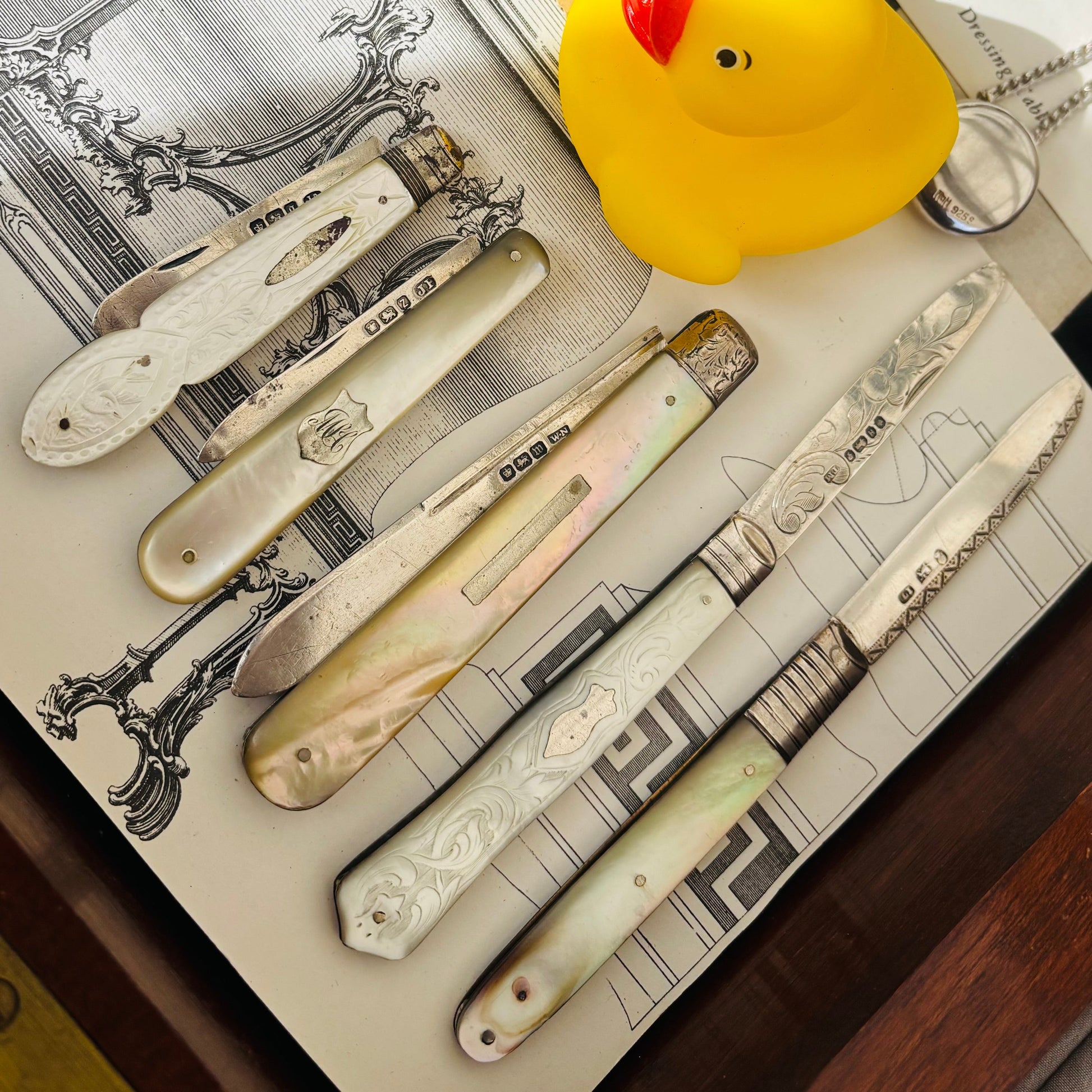 A styled flat-lay of several decorative antique folding fruit knives in silver and mother of pearl, arranged on a vintage-style printed card alongside a silver pocket watch, a small yellow rubber duck, and other antique-themed props.