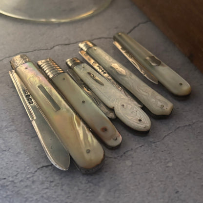 Six antique pocket fruit knives with mother of pearl handles are arranged on a gray stone surface next to a glass stemware base and a wooden box.