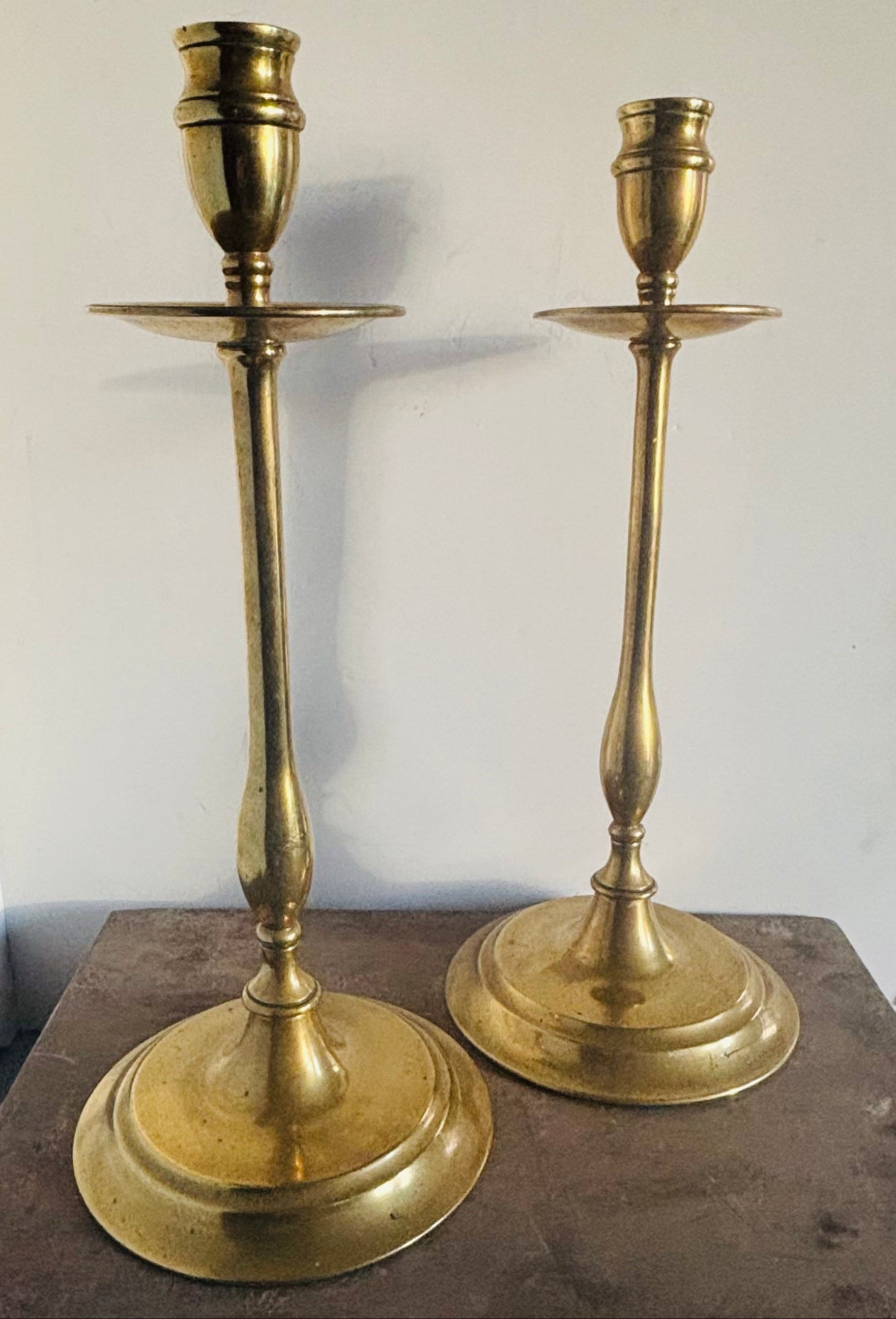A pair of vintage brass candlesticks with a warm gold finish, displayed on a dark wooden surface against a plain off-white wall.