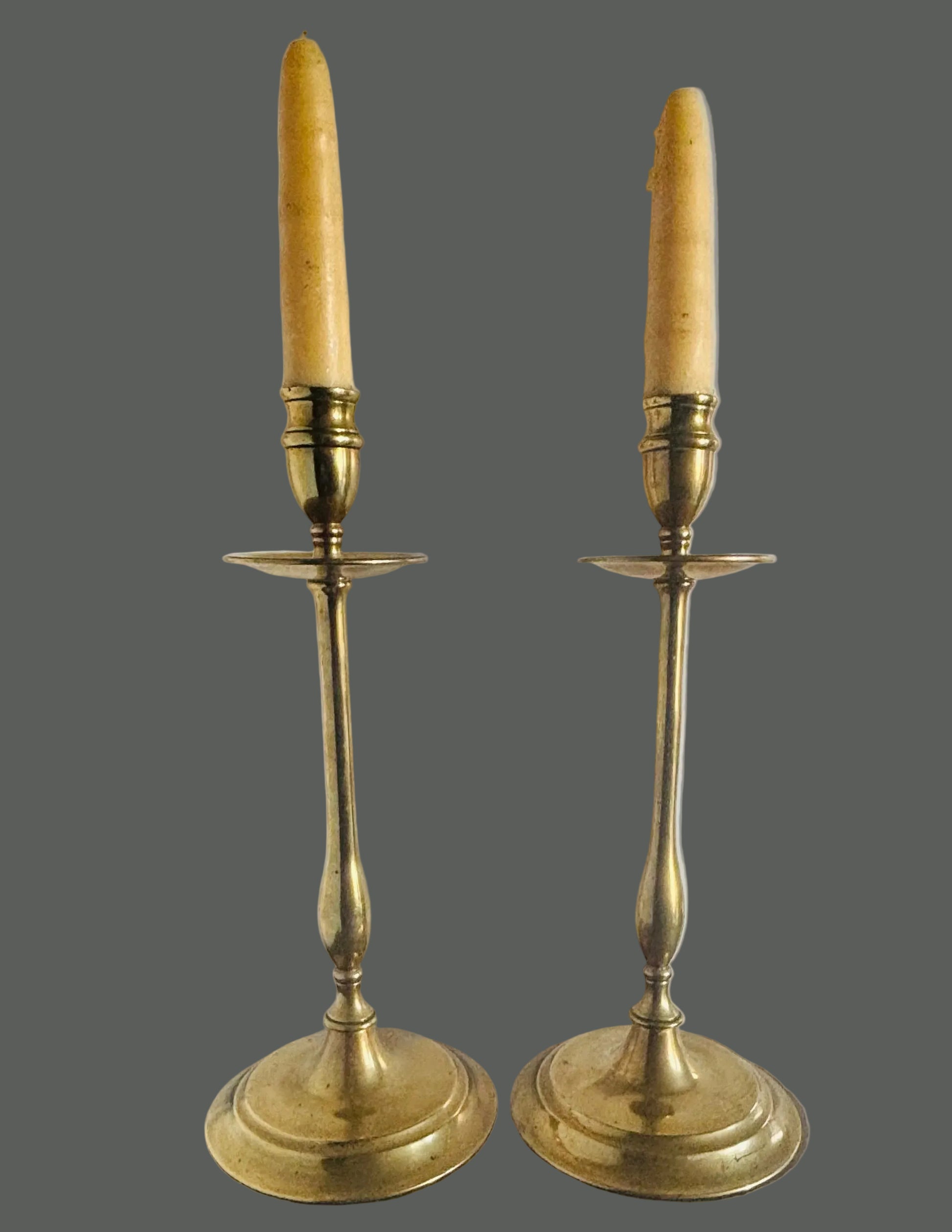A pair of vintage brass candlestick holders with cream-colored taper candles, displayed on a dark wooden surface against a plain white wall.