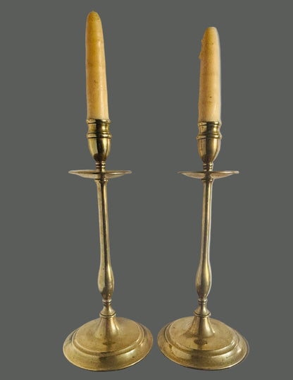 A pair of vintage brass candlestick holders with cream-colored taper candles, displayed on a dark wooden surface against a plain white wall.