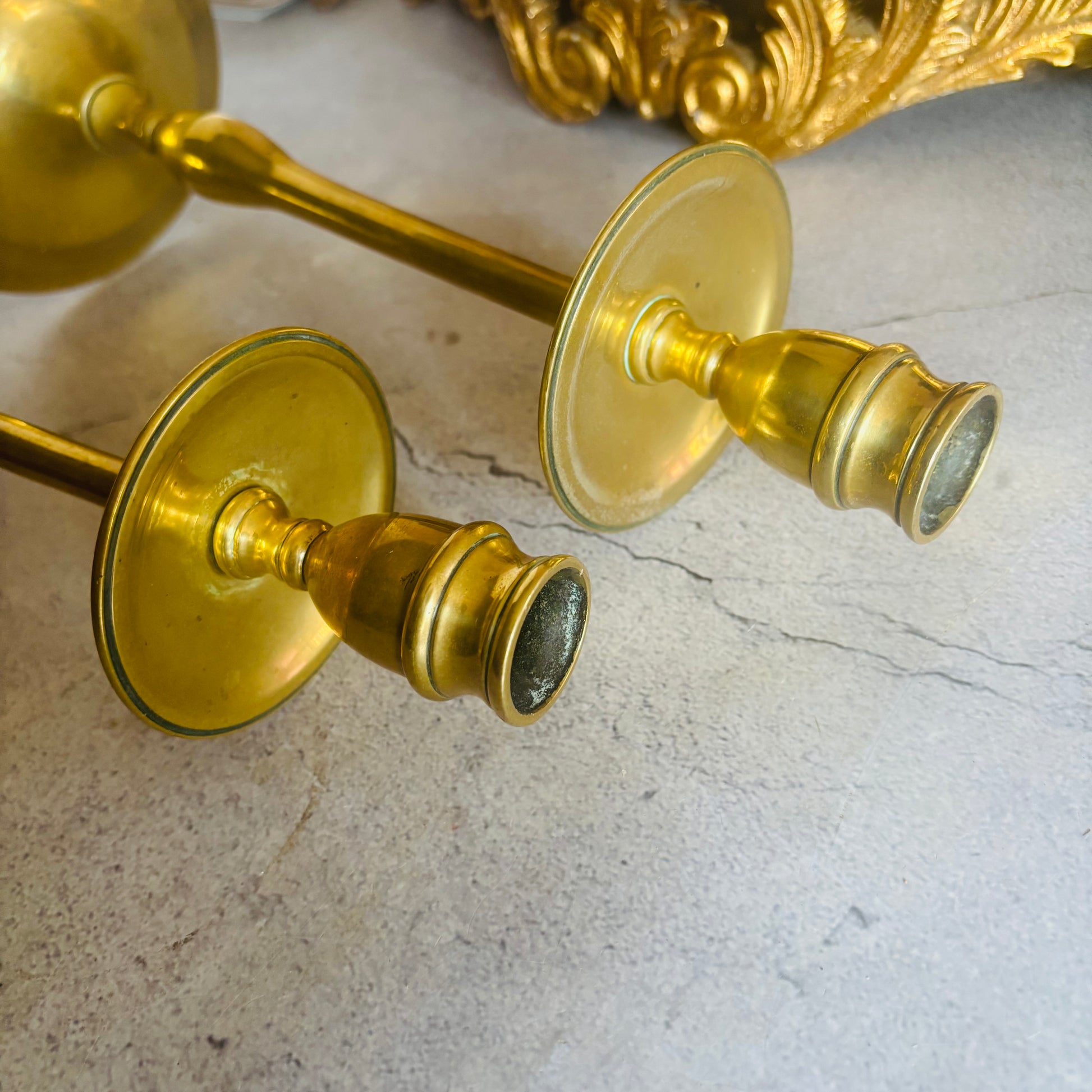 A pair of vintage brass candlesticks with round disc-shaped bases, resting on a light gray stone surface with a decorative gold object in the background.