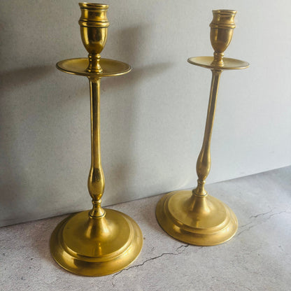 A pair of polished brass candlesticks with a warm gold finish, standing on a light gray textured surface against a neutral gray background.