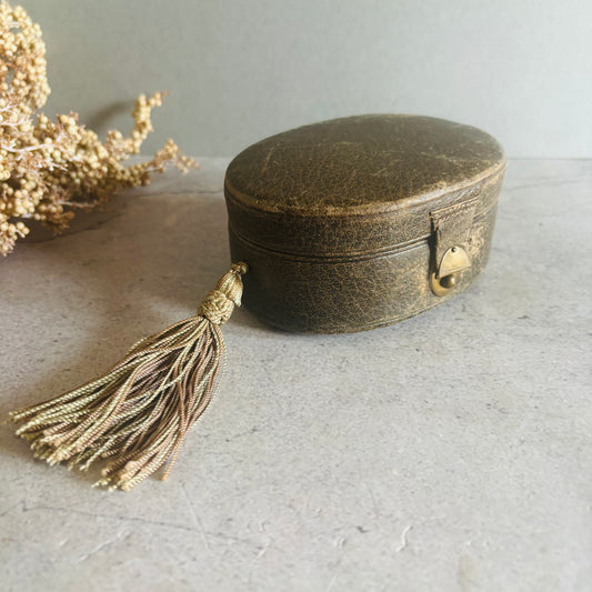 A small oval vintage jewelry box with a distressed dark brown leather-like finish, a gold-tone clasp, and a long braided tassel, photographed on a light gray surface with dried flowers in the background.