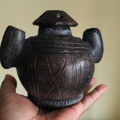 Hand holding a dark brown ceramic pot with decorative patterns against a beige background