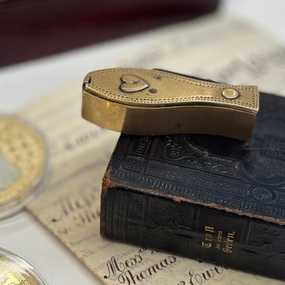 Antique Brass Dutch Coffin Puzzle Snuff Box with a heart-shaped emblem on the cover, displayed on top of an old black leather-bound book over aged paper.