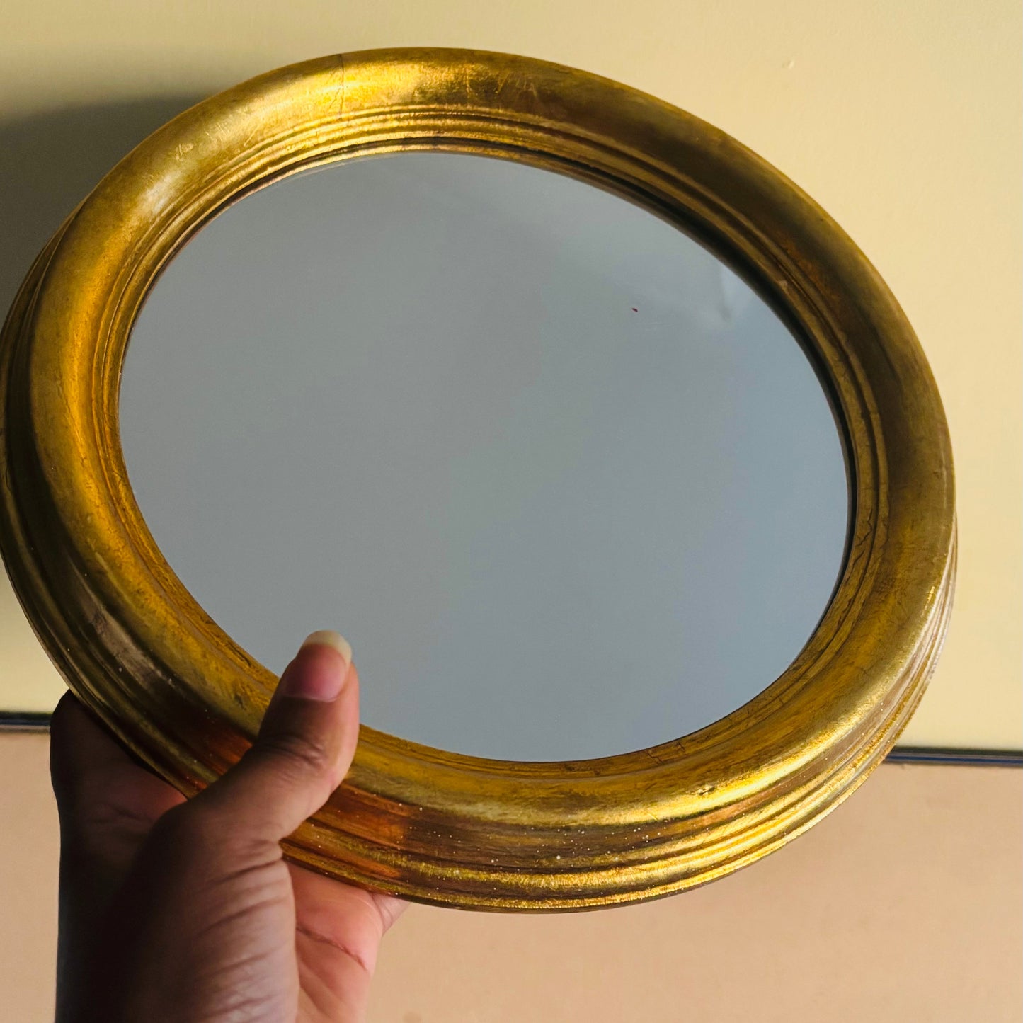 Round gold-framed mirror held by a hand against a neutral background