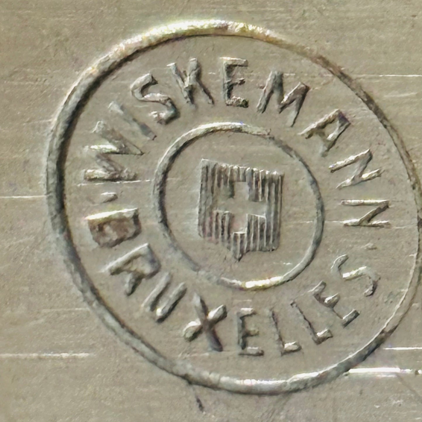 Close-up of a circular stamped logo on a brushed metallic surface, reading 'WISKEMANN BRUXELLES' around a central emblem.