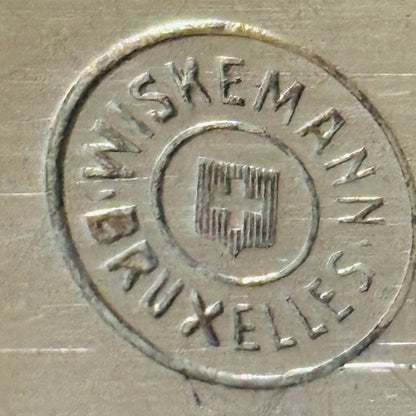Close-up of a circular stamped logo on a brushed metallic surface, reading 'WISKEMANN BRUXELLES' around a central emblem.