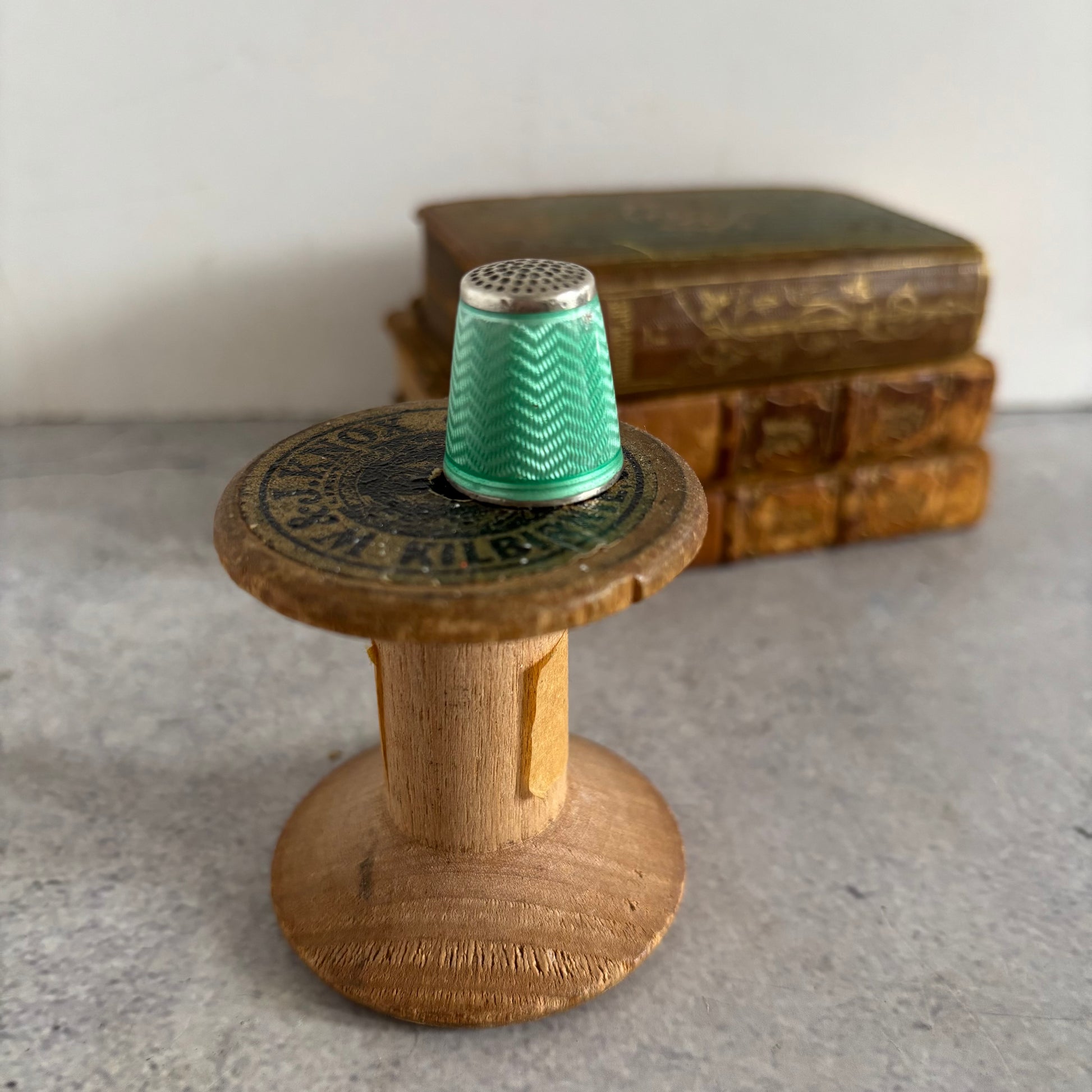Wooden spool with a green thimble on top, placed on a textured surface with books in the background.
