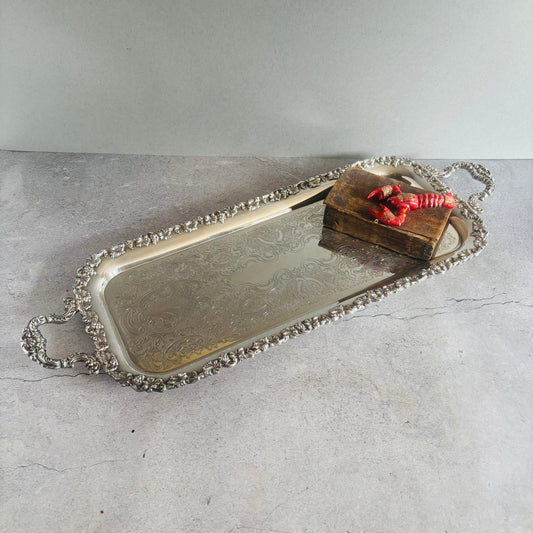 An ornate rectangular silver serving tray with decorative handles and a beaded border, shown on a light gray stone surface with a small leather bible red lobster as styling props.
