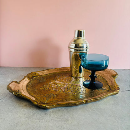 Decorative vintage florentine tray with a silver cocktail shaker and blue glass on a pink background