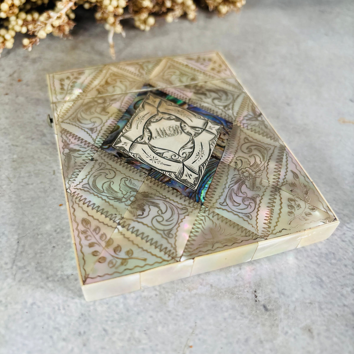 Mother Of Pearl And Sterling Silver Calling Card Case label reading 'MB', placed on a light gray surface with dried flowers in the background.