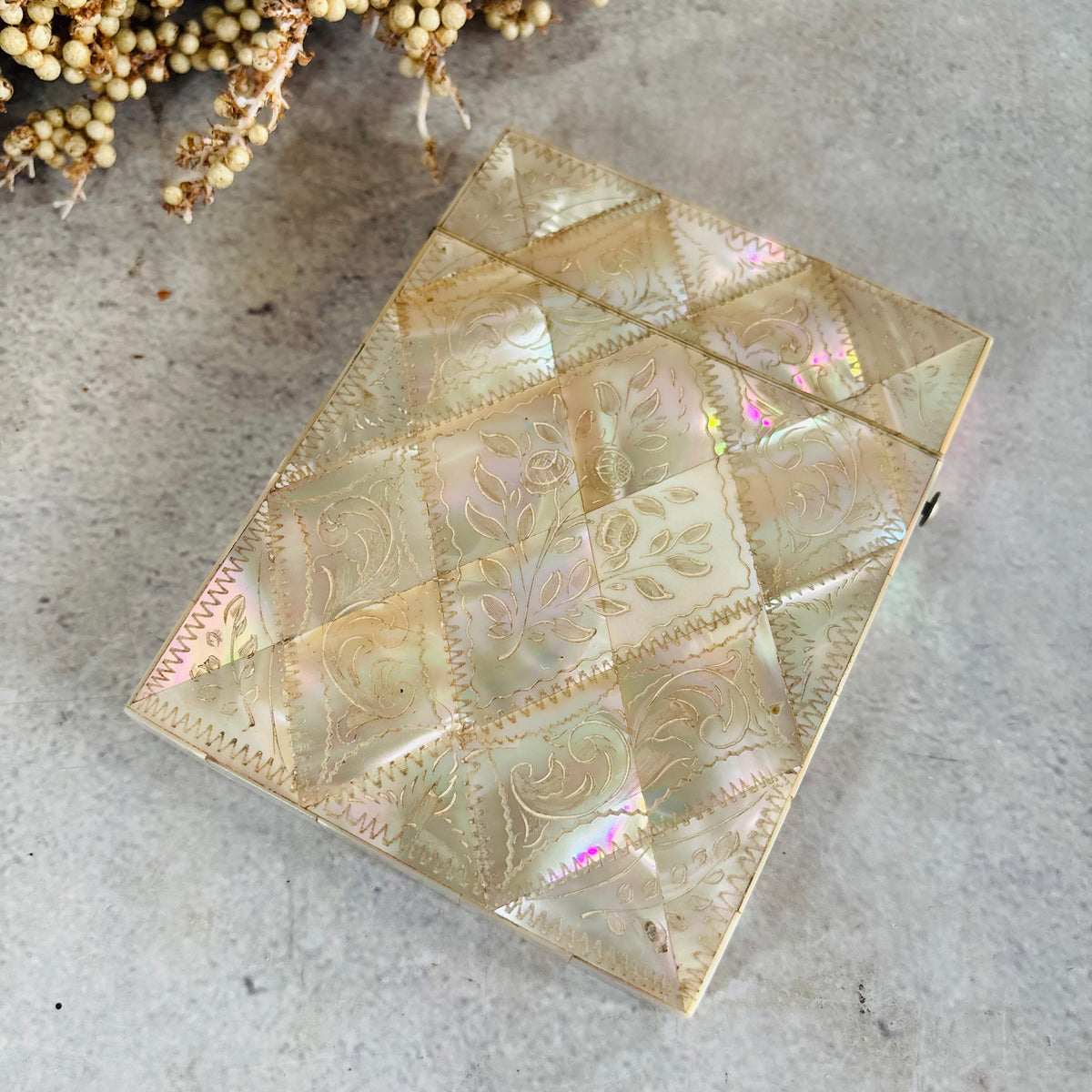 A rectangular iridescent shell or mother-of-pearl card case with engraved floral patterns and geometric paneling, photographed on a light gray textured surface with dried botanicals in the corner.