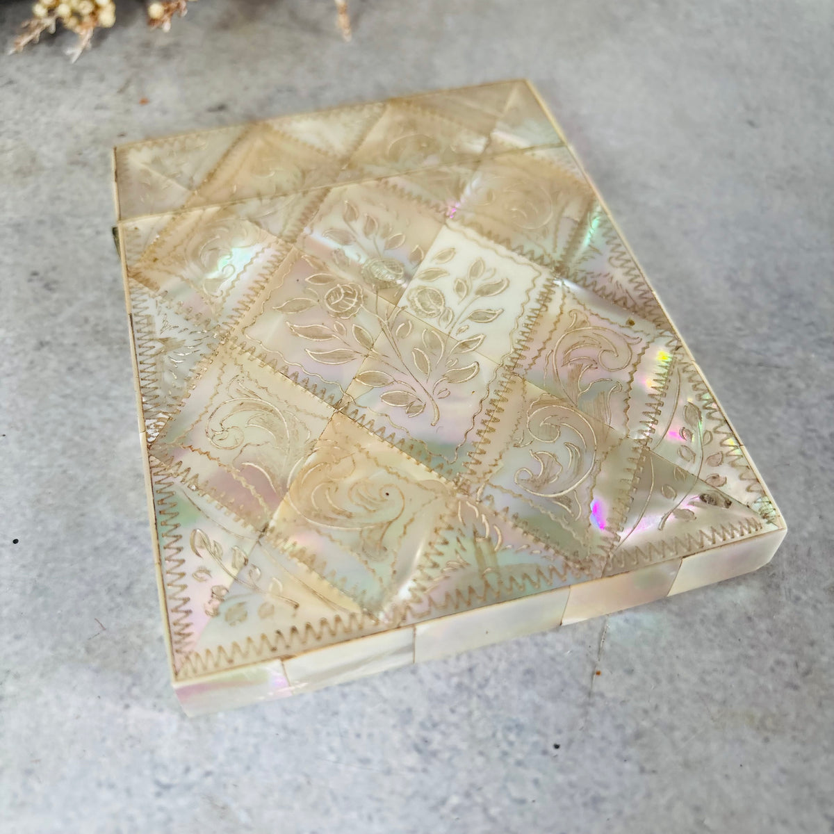 A vintage iridescent shell or mother-of-pearl card case with a diamond quilted pattern and floral engravings, resting on a grey textured surface.