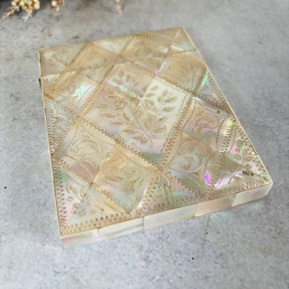 A vintage iridescent shell or mother-of-pearl card case with a diamond quilted pattern and floral engravings, resting on a grey textured surface.