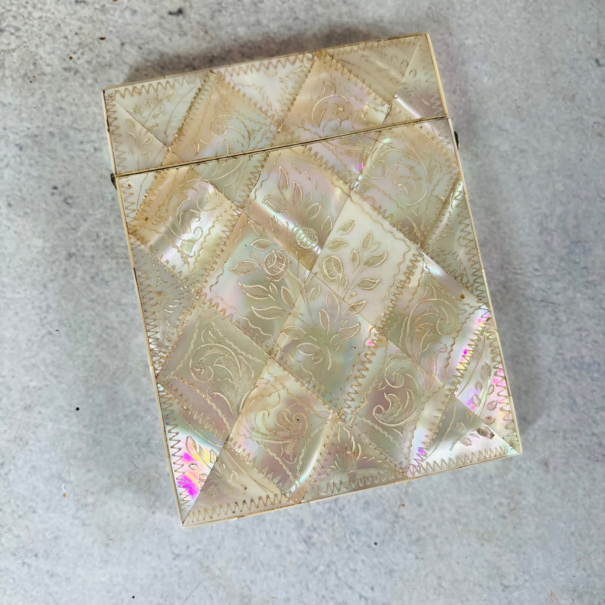 A vintage iridescent shell-covered card case with a diamond quilted pattern and embossed floral motifs, photographed on a textured gray surface.