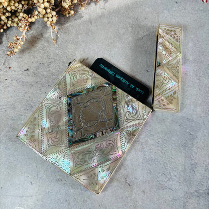 An iridescent mother of pearl card case with embossed geometric and floral patterns, shown open with a business card inside. The compact rests on a textured gray surface with dried flowers in the top-left corner.