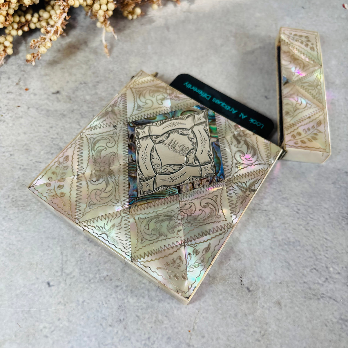 A square iridescent card case with ornate western-style floral patterns in mother of pearl iridescent tones, shown open with a card insert. The case is photographed on a light textured surface with dried flowers in the corner.