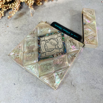 A square iridescent card case with ornate western-style floral patterns in mother of pearl iridescent tones, shown open with a card insert. The case is photographed on a light textured surface with dried flowers in the corner.
