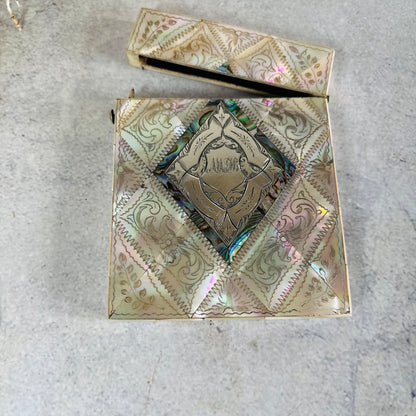 An ornate antique card case with iridescent mother-of-pearl detailing and a central shield emblem, shown open with its lid resting above on a light concrete surface.