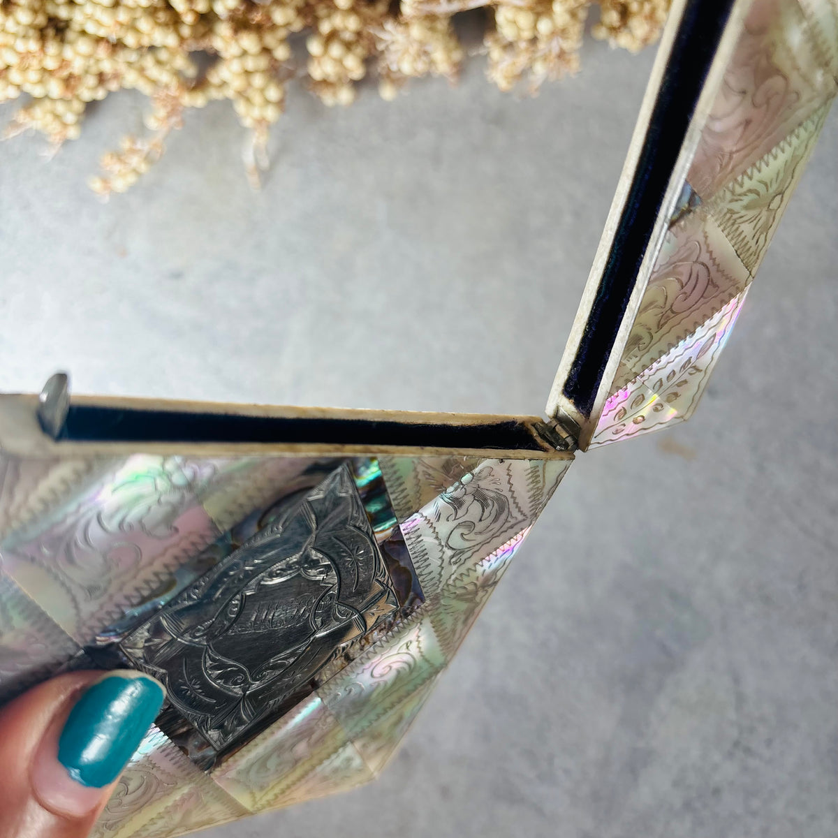 A close-up of a hand with teal nail polish holding open a decorative box lined with iridescent card case, photographed against a light gray surface with dried flowers in the background.