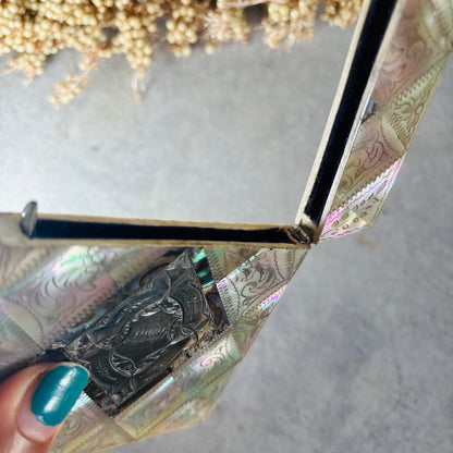 A close-up of a hand with teal nail polish holding open a decorative box lined with iridescent card case, photographed against a light gray surface with dried flowers in the background.