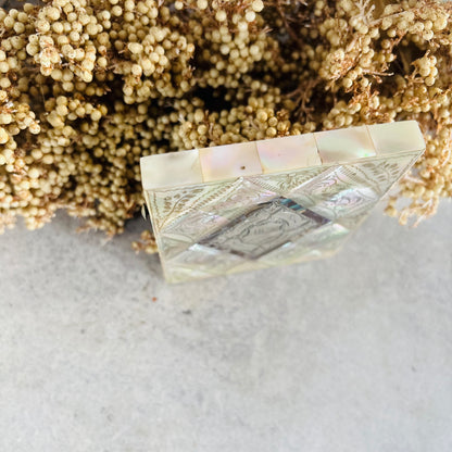 A square decorative box with iridescent mother-of-pearl inlay and a, styled on a light gray surface with dried beige flowers in the background.