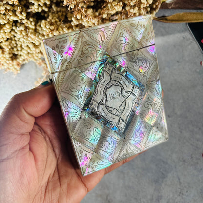 A hand holding a square mother of pearl card case with engraved ornate patterns and a central metallic emblem, photographed against a gray concrete-like surface with dried flowers in the background.