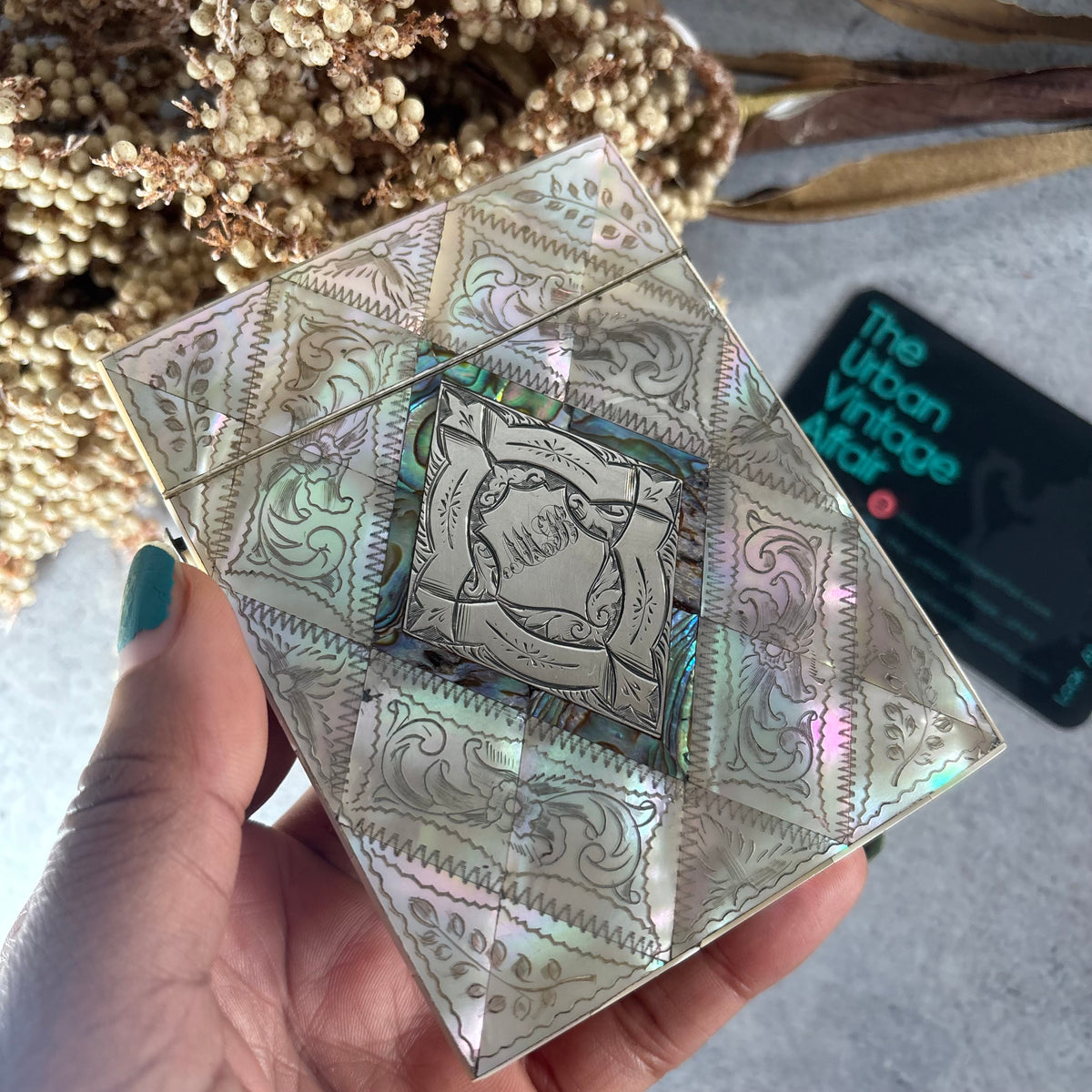 A hand with teal nail polish holds a square, iridescent card case with ornate embossed patterns. A black card reading 'The Urban Vintage Made' is visible in the background along with dried flowers on a gray surface.
