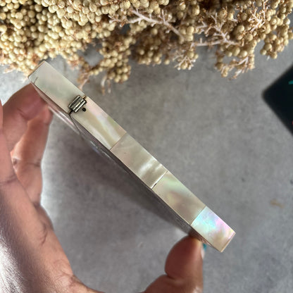 A close-up of a hand holding a small rectangular compact case with iridescent mother-of-pearl style panels and a silver-tone hinge, against a gray surface with dried flowers in the background.