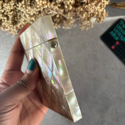 A hand with teal nail polish holds a small rectangular iridescent shell case decorated with a paisley pattern. The case has a silver metal screw cap and is photographed against a gray concrete surface with dried flowers in the background.