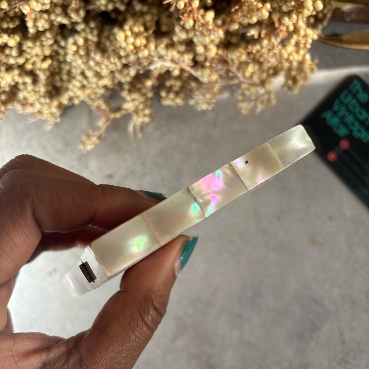 A hand with teal nail polish holds a slim, rectangular card case with iridescent shell-like panels. The background shows a gray surface with dried flowers and a dark card.