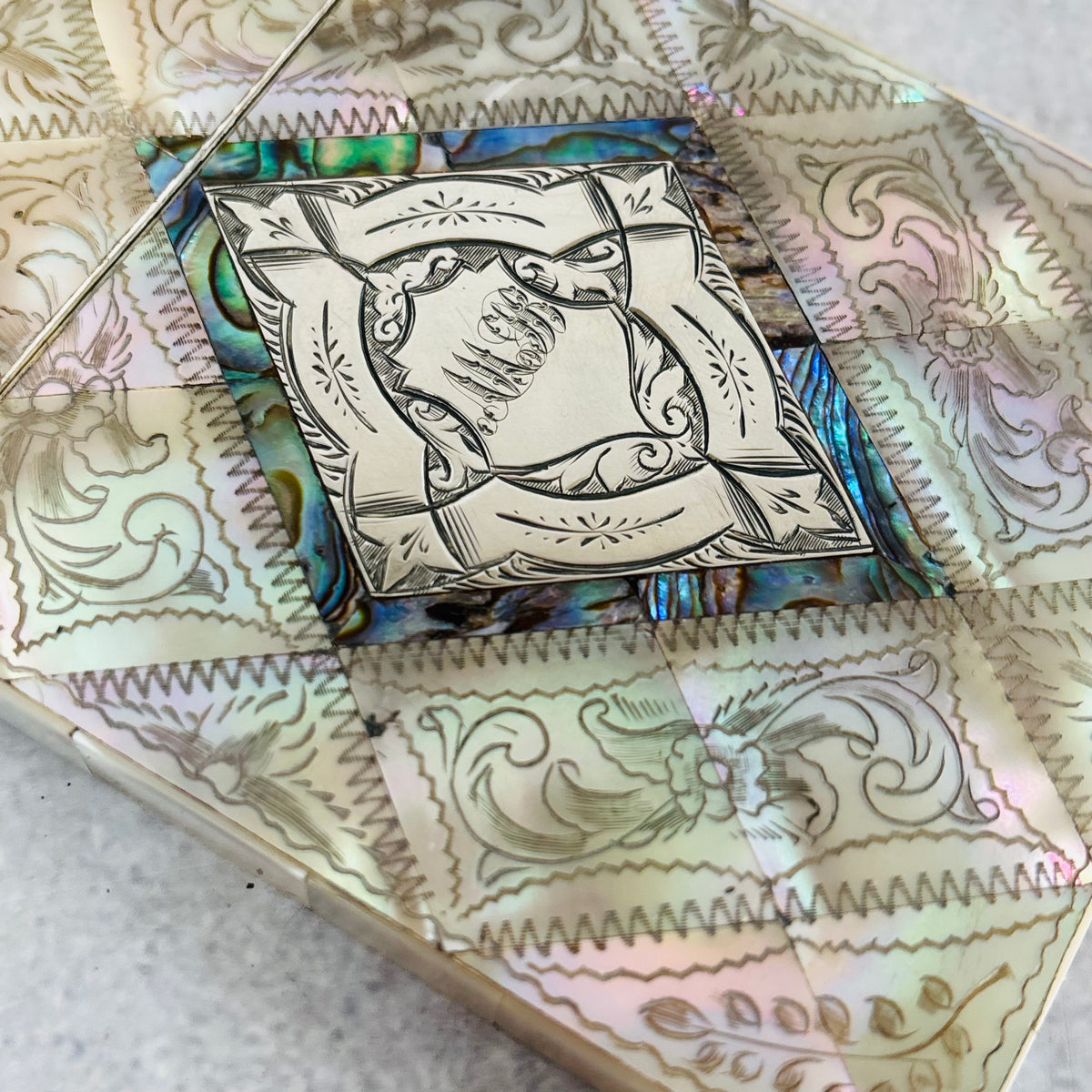 A close-up of Rare Antique Mother Of Pearl And Sterling Silver Calling Card Case  featuring iridescent abalone shell mosaic panels with floral scrollwork and a central silver-toned engraved medallion.