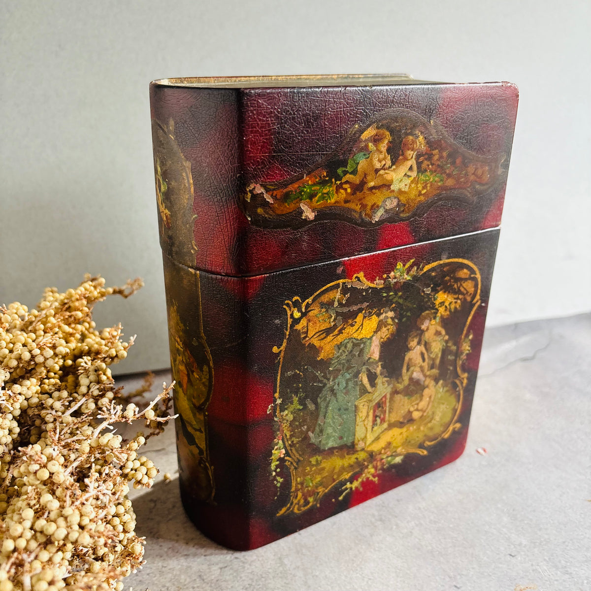 A vintage rectangular lacquered box with a red and black plaid pattern and hand-painted classical figures, shown next to dried flowers on a gray surface.