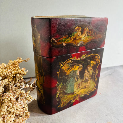 A vintage rectangular lacquered box with a red and black plaid pattern and hand-painted classical figures, shown next to dried flowers on a gray surface.