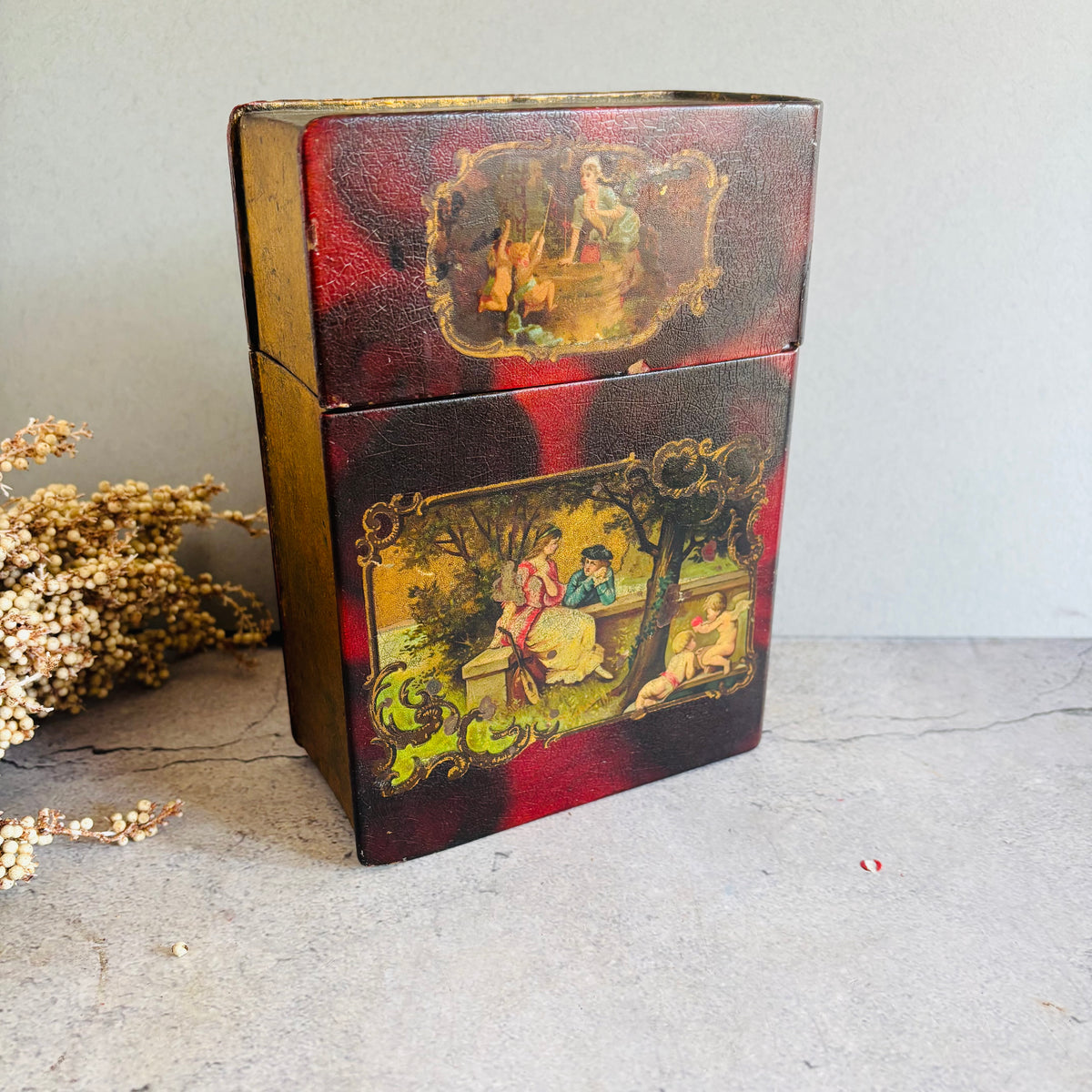 A vintage rectangular metal tin with a hinged lid, decorated with red and gold painted panels featuring classical mythological and pastoral scenes. The tin is photographed on a textured gray surface next to dried white flowers against a neutral gray wall.