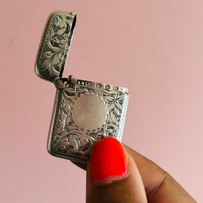 Silver engraved vesta match striker held by a hand with red nail polish against a pink background
