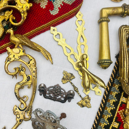 A collection of antique brass and bronze metal hardware, decorative filigree pieces, and ornate brooches arranged on a white surface next to a red and multicolor embroidered textile.