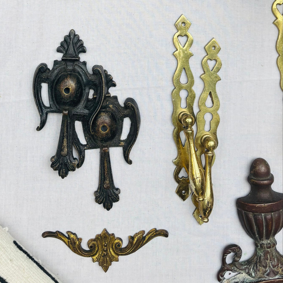 An assortment of vintage ornate metal hardware pieces, including brass and bronze decorative pieces, a dark bronze handle, and a small copper-toned piece, arranged on a white textured fabric background.