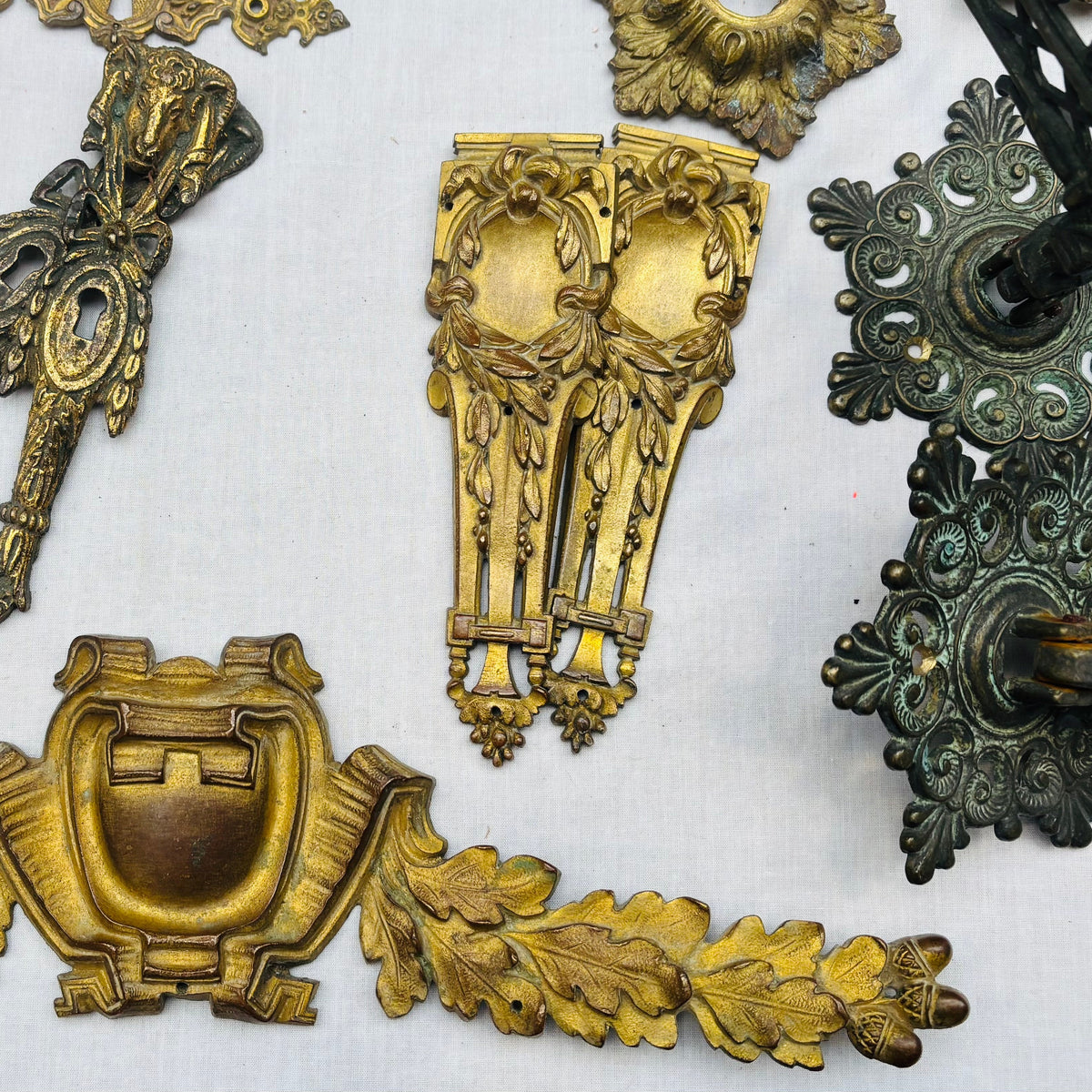 A collection of ornate vintage brass and bronze hardware pieces, including decorative brackets, wall mounts, and sculpted accents, arranged on a white fabric background.