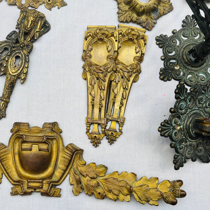 A collection of ornate vintage brass and bronze hardware pieces, including decorative brackets, wall mounts, and sculpted accents, arranged on a white fabric background.
