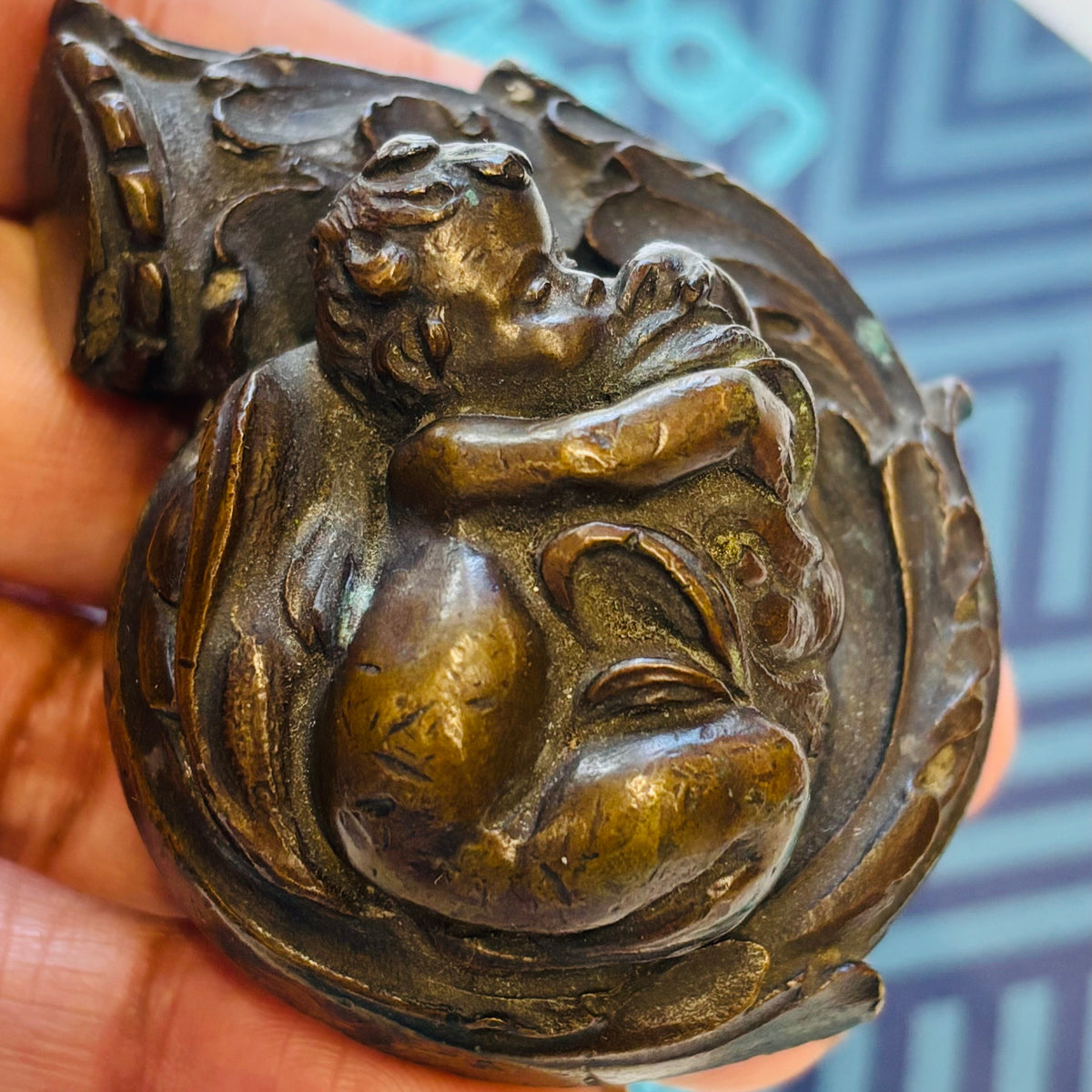 A hand holding a small bronze figurine of a cherub curled up inside a crescent moon, with a blue patterned card in the background.