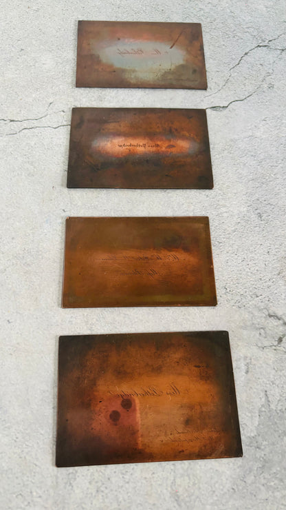Four rectangular copper plates with engraved text and decorative patterns, arranged vertically on a textured light gray concrete surface.