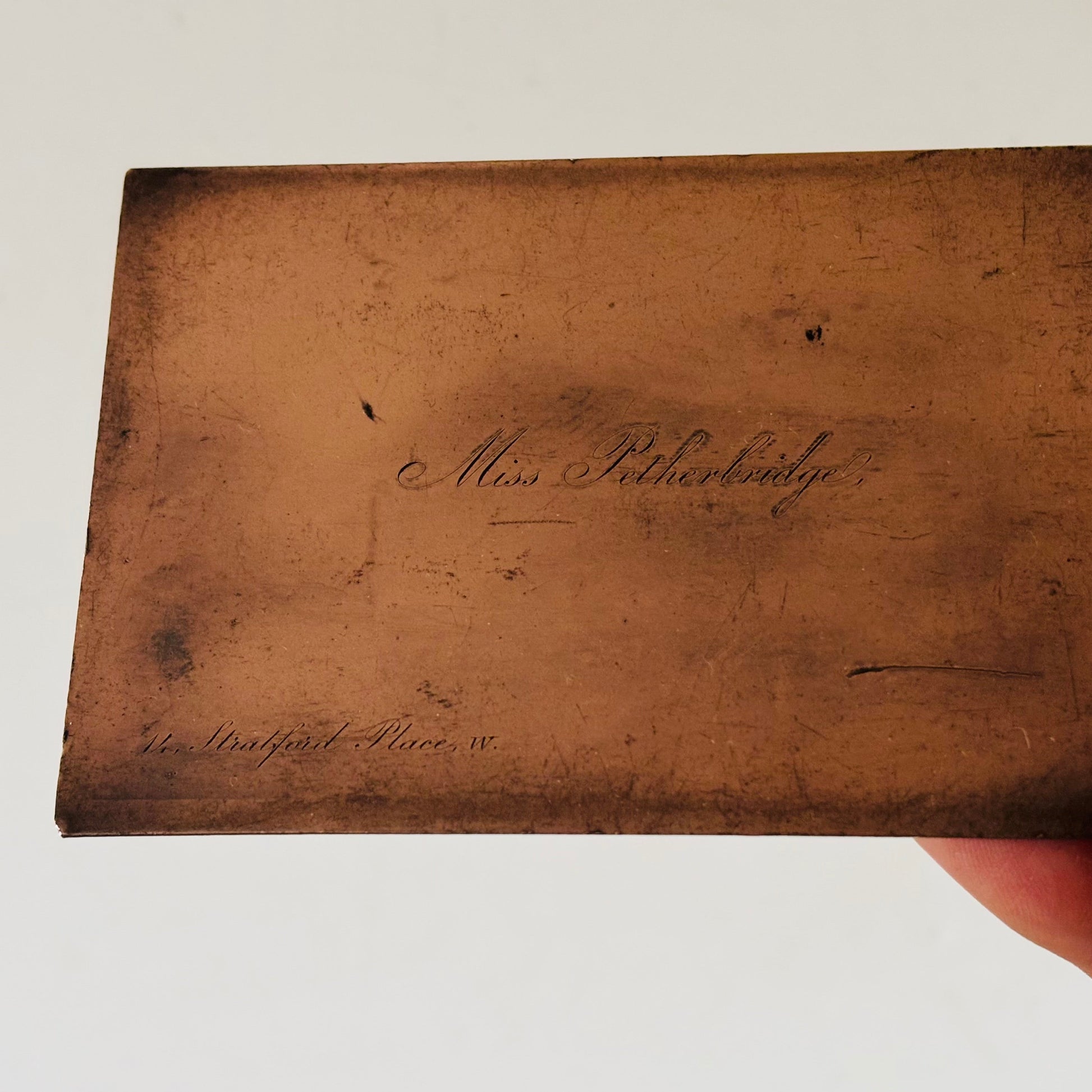 A close-up of a copper printing plate with faint cursive handwriting, held against a plain white background with a finger visible at the bottom right.