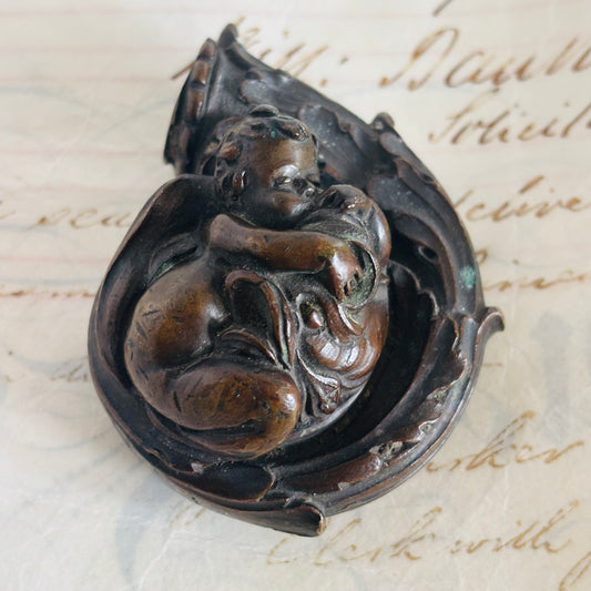 A dark brown Antique Bronze Sculpture of Sleeping Putto displayed on a vintage, handwritten paper background.